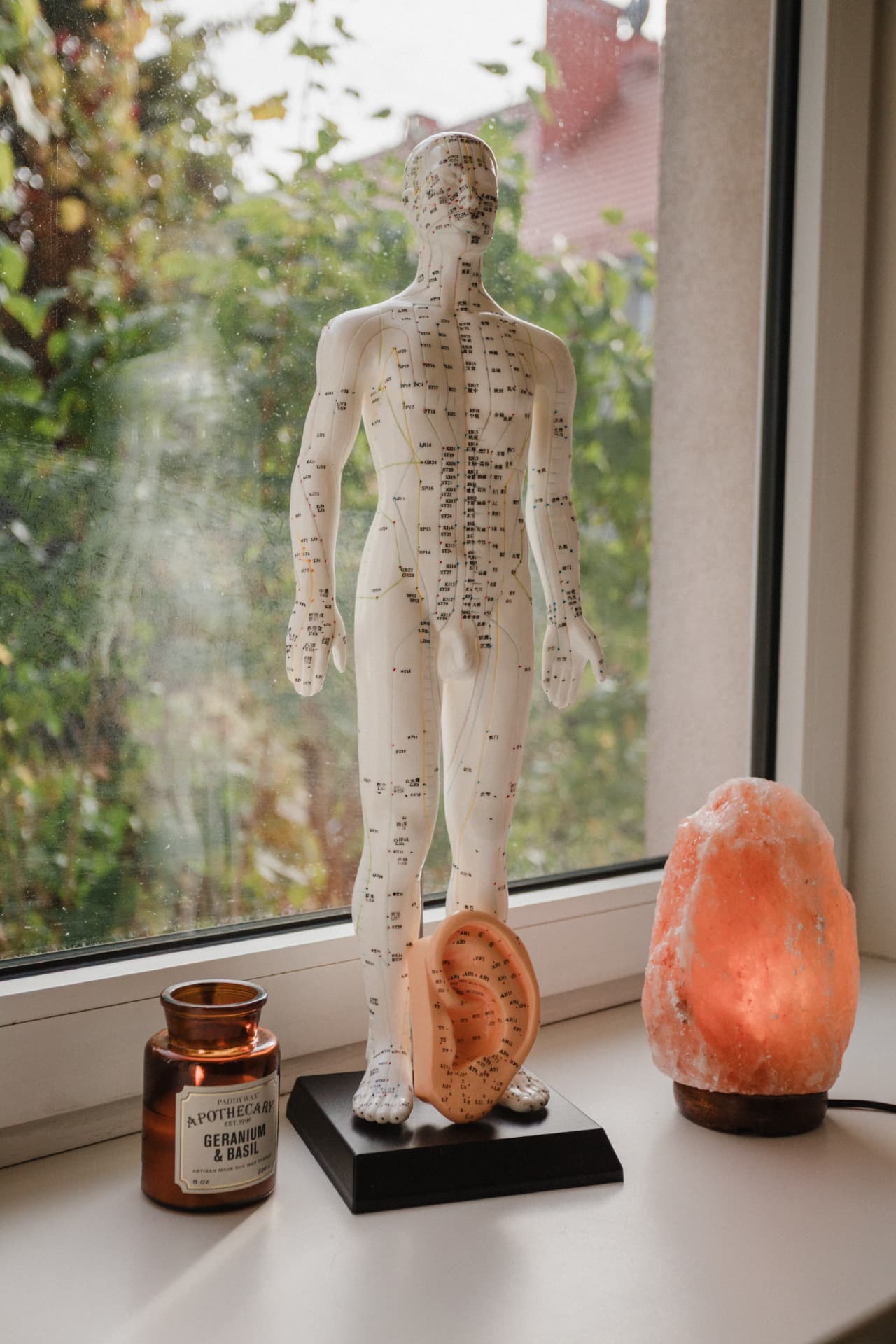 Model anatomiczny akupunktury z zaznaczonymi punktami i model ucha na białym parapecie, obok bursztynowego słoika ze świecą i lampy solnej, za oknem zieleń drzew